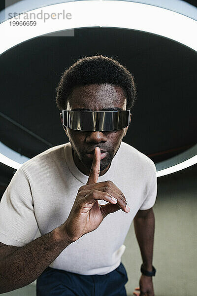 Junger Mann mit futuristischer Cyberbrille unter moderner Ringlampe  der den Zeigefinger auf die Lippen legt