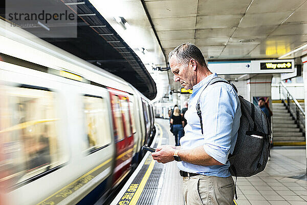 Geschäftsmann benutzt Smartphone und wartet auf dem Bahnsteig auf den Zug