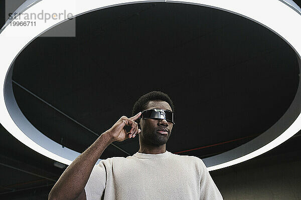 Junger Mann mit futuristischer Cyberbrille steht unter moderner Ringlampe und legt den Finger auf die Schläfen