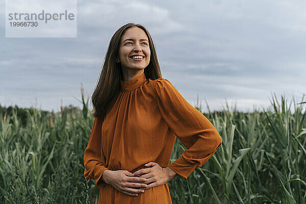 Fröhliche Frau mit den Händen auf der Hüfte steht neben Maispflanzen auf dem Feld