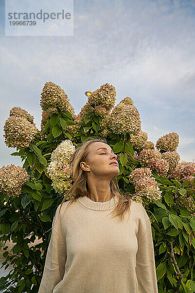 Junge Frau steht mit geschlossenen Augen vor einer blühenden Pflanze unter dem Himmel