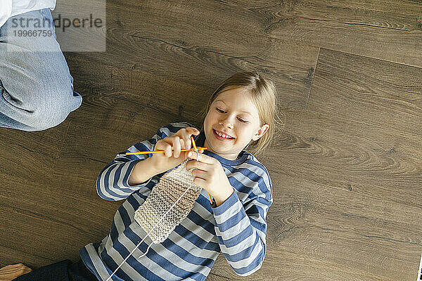 Smiling girl doing knitting lying on wooden floor at home