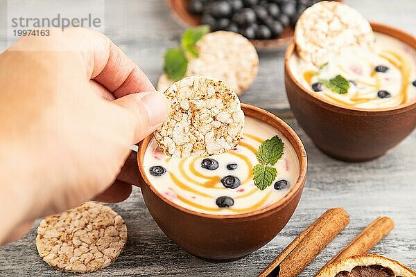 Joghurt mit Heidelbeeren und Karamell in Tonschale auf grauem Holz  Hand taucht Kekse in Joghurt  Seitenansicht  Nahaufnahme  selektiver Fokus