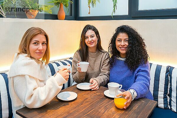 Drei Freundinnen schauen in die Kamera und trinken gemeinsam Kaffee in einer netten Cafeteria