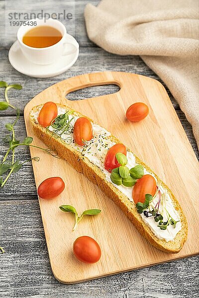 Langes Weißbrot Sandwich mit Frischkäse  Tomaten und Mikrogrün auf grauem Holzhintergrund und Leinenstoff. Seitenansicht  Nahaufnahme  selektiver Fokus