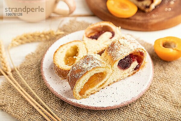 Hausgemachtes süßes Brötchen mit Aprikosenmarmelade und Tasse Kaffee auf weißem Holzhintergrund und Leinenstoff. Seitenansicht  Nahaufnahme  selektiver Fokus