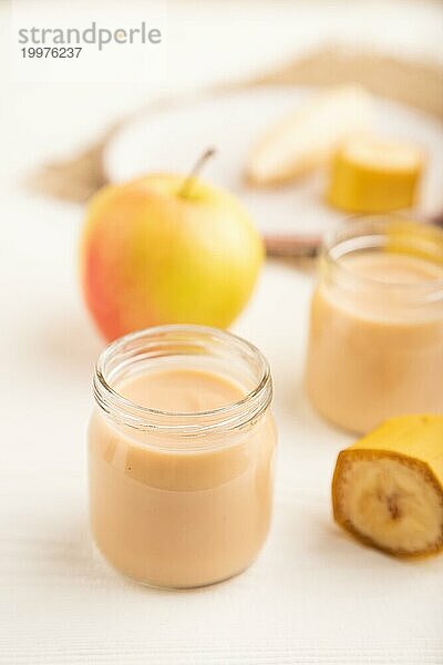 Baby Püree mit Früchten mischen  Apfel  Banane Säuglingsnahrung in Glasgefäß auf weißem Holz Hintergrund. Seitenansicht  Nahaufnahme  selektiver Fokus  künstliche Ernährung Konzept