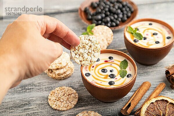 Joghurt mit Heidelbeeren und Karamell in Tonschale auf grauem Holz  Hand taucht Kekse in Joghurt  Seitenansicht  Nahaufnahme  selektiver Fokus