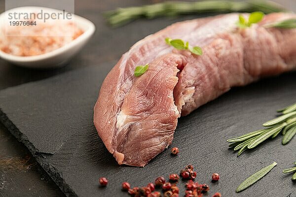 Rohes Schweinefleisch mit Kräutern und Gewürzen auf Schiefer Schneidebrett auf schwarzem Beton Hintergrund. Seitenansicht  Nahaufnahme  selektiver Fokus