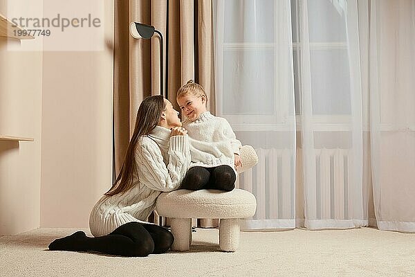 Eine junge Mutter schaut liebevoll auf ihre kleine Tochter  die auf einem weichen Stuhl sitzt