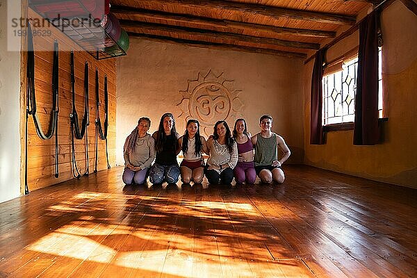 Gruppenporträt einer Gruppe von Menschen  die lächelnd Yoga praktizieren und in die Kamera schauen. Allgemeine Aufnahme  beleuchtet mit natürlichem Licht  das durch die Seitenfenster einfällt