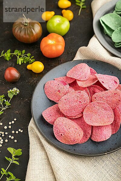 Grüne und rote Kartoffelchips mit Kräutern und Tomaten auf schwarzem Betonhintergrund und Leinenstoff. Seitenansicht  Nahaufnahme  selektiver Fokus