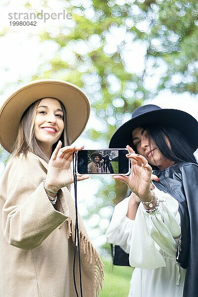 Vertikale Vorderansicht von zwei stilvollen Freundinnen  die beide ein Handy in die Hand nehmen  um ein Selfie zu machen