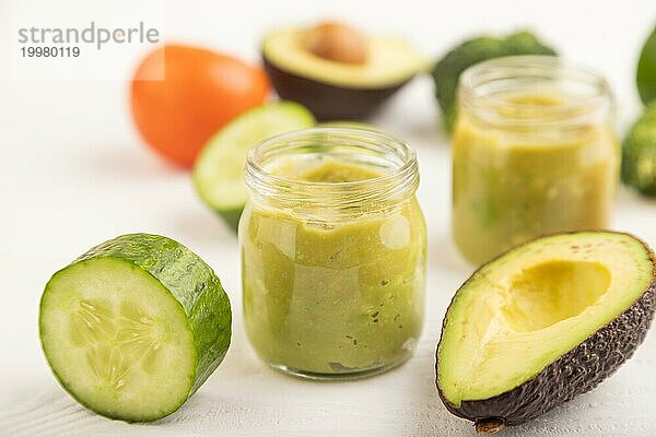 Baby Püree mit Gemüsemischung  Brokkoli  Tomaten  Gurken  Avocado Säuglingsnahrung in Glasgefäß auf weißem Holzhintergrund. Seitenansicht  Nahaufnahme  selektiver Fokus  Konzept der künstlichen Ernährung