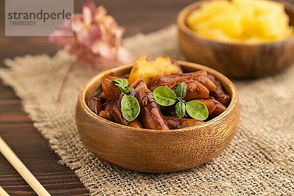 Tteokbokki oder Topokki  gebratener Reiskuchen am Stiel  beliebtes koreanisches Straßenessen mit würziger Jjajang Soße und Ananas auf braunem Holzhintergrund und Leinenstoff. Seitenansicht  selektiver Fokus