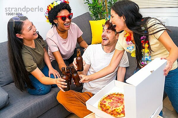 Multiethnische Freunde stoßen mit Bier an  essen Pizza und feiern auf dem Sofa sitzend ihren Geburtstag