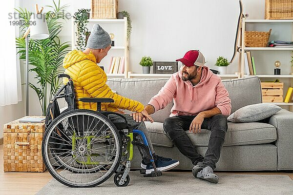 Mann unterstützt einen behinderten Freund im Rollstuhl  während er zu Hause auf dem Sofa sitzt