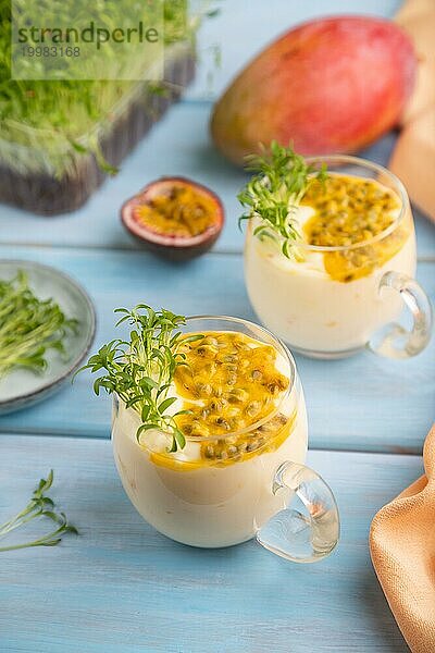 Mango Joghurt mit Passionsfrucht und Koriander Mikrogrün im Glas auf blauem Holzhintergrund mit orangefarbenem Leinenstoff. Seitenansicht  Nahaufnahme  selektiver Fokus