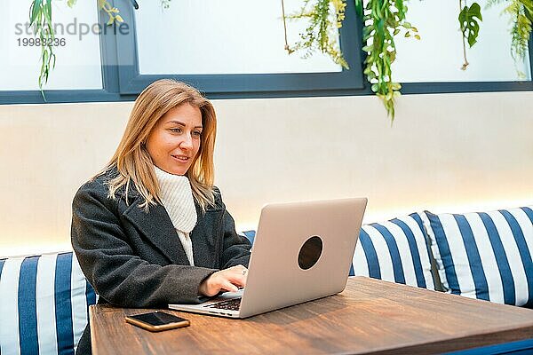 Elegante erwachsene blonde Frau arbeitet mit Laptop in einer bunten Cafeteria sitzen