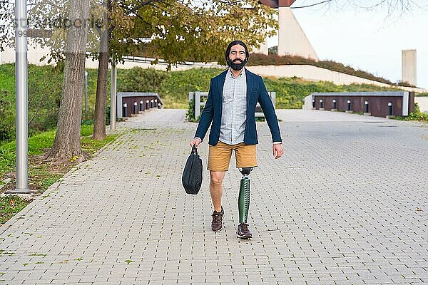 Frontalansicht eines Geschäftsmannes mit Beinprothese  der eine Laptoptasche durch einen Stadtpark trägt