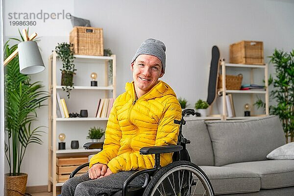 Porträt eines fröhlichen behinderten Mannes im Rollstuhl  der in die Kamera lächelt  im Wohnzimmer zu Hause