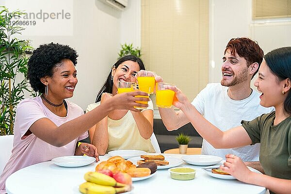 Fröhliche junge multiethnische Freunde stoßen beim Frühstück zu Hause mit Orangensaft an