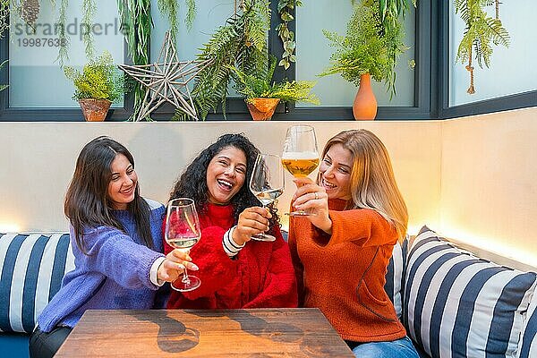 Horizontales Foto von drei Freundinnen  die in einer modernen bunten Cafeteria sitzen und mit Weißwein anstoßen