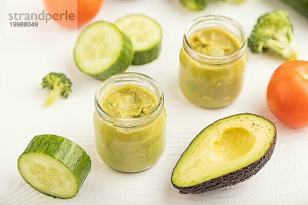 Baby Püree mit Gemüsemischung  Brokkoli  Tomaten  Gurken  Avocado Säuglingsnahrung in Glasgefäß auf weißem Holzhintergrund. Seitenansicht  Nahaufnahme  selektiver Fokus  Konzept der künstlichen Ernährung