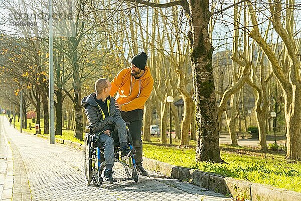 Mann  der den Rollstuhl eines behinderten Freundes auf dem Weg durch einen Park schiebt