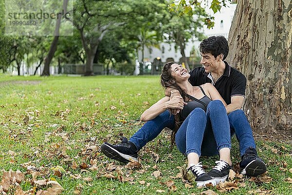 Paar sitzt in einem Park und lehnt sich an einen Baum. Glückliches Liebespaar lachend
