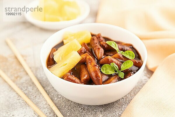 Tteokbokki oder Topokki  gebratener Reiskuchen am Stiel  beliebtes koreanisches Straßenessen mit würziger Jjajang Sauce und Ananas auf grauem Betonhintergrund und orangefarbenem Textil. Seitenansicht  Nahaufnahme  selektiver Fokus