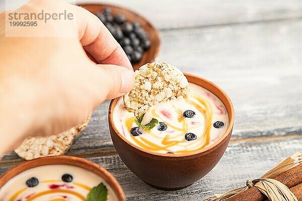 Joghurt mit Heidelbeeren und Karamell in Tonschale auf grauem Holz  Hand taucht Kekse in Joghurt  Seitenansicht  Nahaufnahme  selektiver Fokus