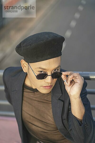 Latino schwuler Männchen mit Makeup und modischem Hut posiert auf einer Brücke