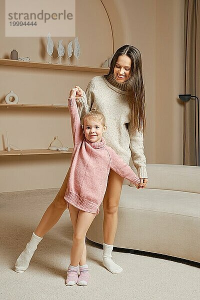 Eine fröhliche Mutter und ihre kleine Tochter tanzen zusammen in einem gemütlichen Wohnzimmer. Die Tochter trägt einen rosa Pullover  während die Mutter einen grauen Pullover trägt