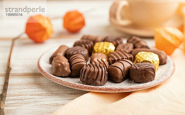 Schokoladenbonbons mit Kaffeetasse und Physalisblüten auf weißem Holzhintergrund und orangefarbenem Textil. Seitenansicht  Nahaufnahme  selektiver Fokus