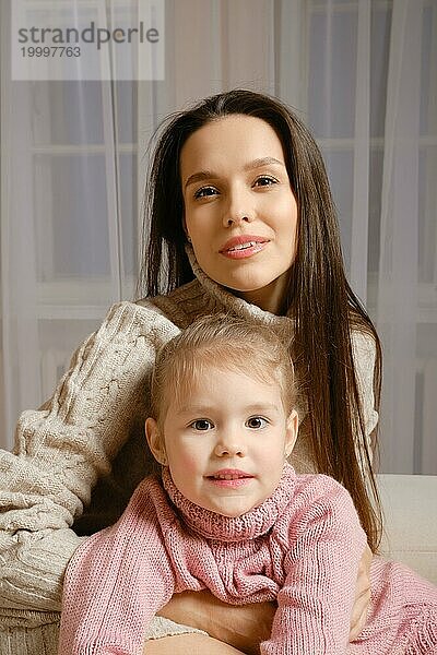 Eine schöne und liebevolle Mutter und eine süße Tochter sitzen in gestrickten Pullovern zusammen im Wohnzimmer