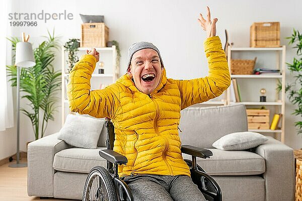 Porträt eines glücklichen behinderten Mannes im Rollstuhl  der vor Freude die Hände hebt und im Wohnzimmer zu Hause feiert