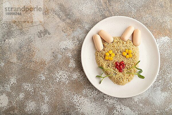 Lustig gemischter Quinoa Brei  Mais  Granatapfelkerne und kleine Würstchen in Form eines Katzengesichts auf braunem Betonhintergrund. Draufsicht  flat lay  copy space. Essen für Kinder  gesundes Essen Konzept