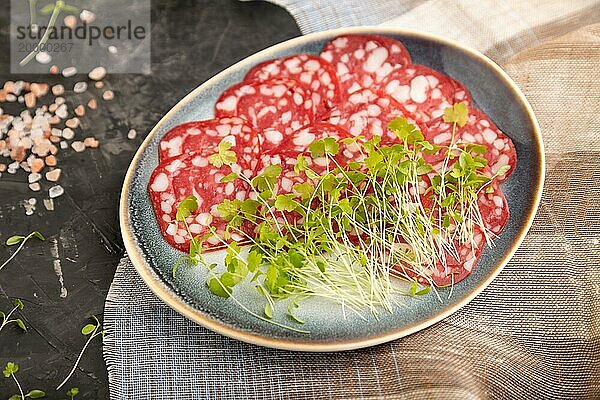 Scheiben von geräucherter Cervelatwurst mit Spinat  Salz und Pfeffer auf schwarzem Betonhintergrund und beigem Textil. Seitenansicht  Nahaufnahme  selektiver Fokus