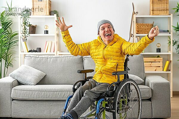 Glücklicher und ausdrucksstarker behinderter Mann im Rollstuhl  der zu Hause die Hände vor Freude hebt