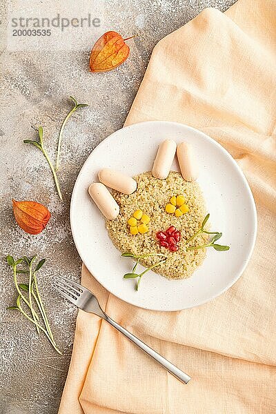 Lustig gemischter Quinoa Brei  süßer Mais  Granatapfelkerne und kleine Würstchen in Form eines Katzengesichts auf braunem Betonhintergrund und orangefarbenem Textil. Draufsicht  flach gelegt. Essen für Kinder  gesundes Essen Konzept