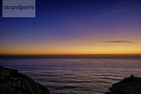 Schöne ruhige Blick auf den Ozean bei Sonnenuntergang  mit einem Farbverlauf von orange bis lila  dunkle hohe Klippen und Mondsichel in den Himmel. Bunte klaren Himmel in der Dämmerung  Horizont über Wasser  ruhige Gewässer. Meereslandschaft im Hintergrund