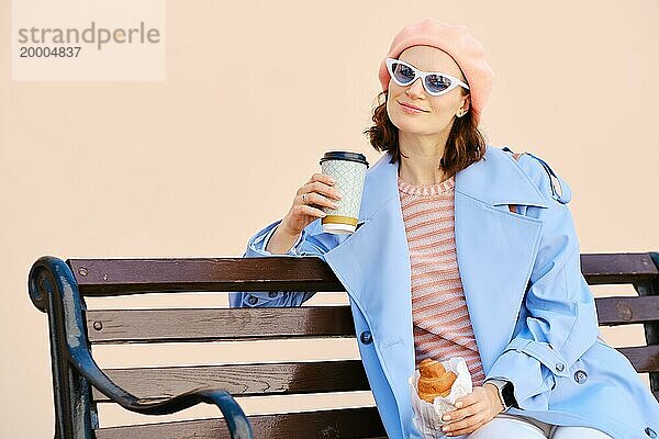 Zufriedene Frau setzt sich auf eine Bank  um Kaffee zu trinken und ein Croissant zu essen