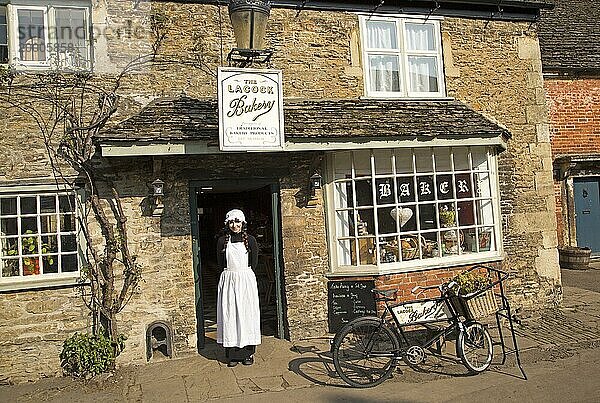 Junge Ladenbesitzerin in altmodischer traditioneller Kleidung vor dem Dorfbäckerladen  Lacock  Wiltshire  England  UK