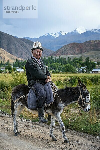Mann in traditioneller Kleidung reitet auf einem Esel vor einer Bergkulisse  Kirgistan  Asien