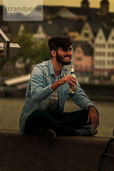 Ein Mann sitzt am Flussufer und genießt ein Getränk bei abendlicher Stimmung  Hohenzollernbrücke  Köln Deutz  Nordrhein-Westfalen  Deutschland  Europa