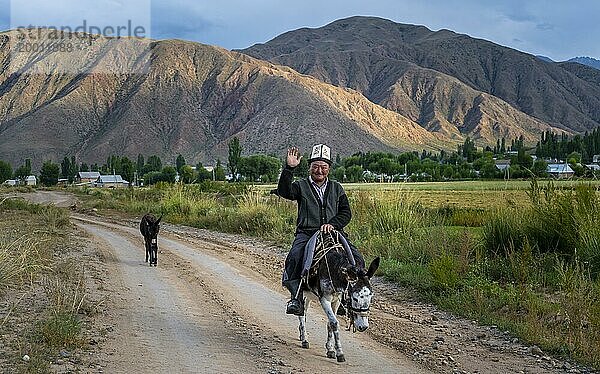 Mann in traditioneller kirgisischer Kleidung reitet auf einem Esel entlang einer Dorfstraße und grüßt freundlich  Kirgistan  Asien