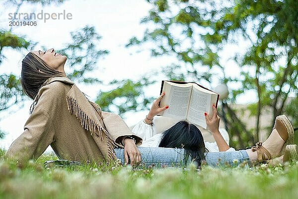 Seitenansicht von zwei entspannten Freundinnen  die einen Tag zusammen im Park genießen  während sie frische Luft schnappen und ein Buch lesen