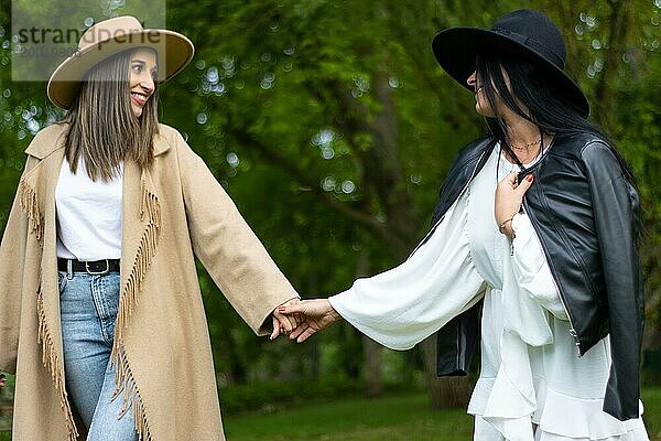 Vorderansicht eines stilvollen lesbischen Paares mit Hut  das sich an den Händen hält und im Park spazieren geht  während es sich gegenseitig anschaut