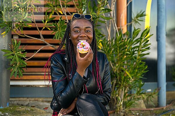 Nettes und glückliches afrikanisches Mädchen mit Lederjacke und Sonnenbrille  das einen Donut isst und allein auf der Straße sitzt